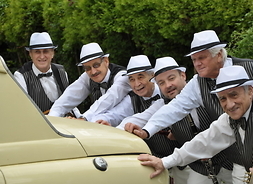 Grupa muzyków, jeden trzyma trąbkę, inny klarnet, opartych o zabytkowy samochód, ubranych w letnie kapelusze, koszule z kamizelką i muszką