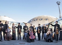 Grupa muzyków w strojach koncertowych z instrumentami na dziedzińcu przed budynkiem o futurystycznych kształtach – kopuły połączone oszklonymi przejściami