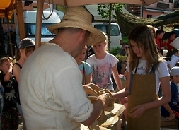 Dwoje dzieci patrzy, a jedna dziewczynka próbuje toczyć wazonik z gliny przy garncarzu na stoisku jarmarcznym. W tle kolejne stoiska