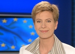 Zdjęcie portretowe dziennikarki w białym żakiecie i koszuli w paski w studiu telewizyjnym. Na ścianie ekran wyświetlający flagę Unii Europejskiej