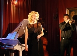 Artystka w wieczorowej sukience, za nią fortepian, obok ubrany w dżinsy i koszulę muzyk z trąbką przy drugim mikrofonie
