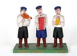 Na podstawce trzy figury muzyków w strojach ludowych (m.in. wysokie buty i czapki), z instrumentami: skrzypcami, akordeonem i czynelami