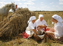 Dwie żniwiarki w strojach ludowych siedzą nad koszem i dzielą wyjęty z niego chleb. Za nimi styg zboża, kolejna kobieta prowadząca z uzdę konia i pole pełne stygów