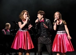 Na zdjęciu sześciu muzyków ubranych w eleganckie czarno-różowe stroje. Grają na fortepianie, perkusji i kontrabasie oraz śpiewają