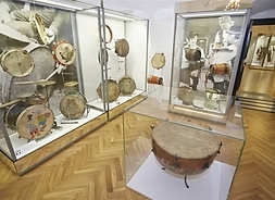 Kolekcja różnego rodzaju bębnów, kotłów, perkusji i tamburynów rozmieszczonych w szklanych gablotach sali w muzeum z podłogą z drewnianej klepki