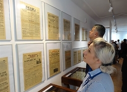 Kobieta i mężczyzna stojący przed zawieszonymi na ścianie ramkami, w których widać pierwsze strony gazet za szkłem, m.in. „Kurjer Poranny” i „Wieczór! Kurjer”