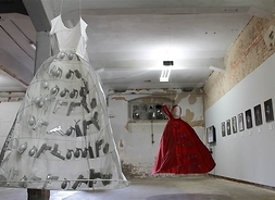 Dwie półprzezroczyste suknie z zaszytymi w nich licznymi pistoletami i granatami. Na ścianach widoczne stare portrety kobiet