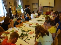 Grupa 13 dzieci i dwoje dorosłych siedzą przy stole, na którym widać materiały malarskie (pudełka z farbami, kartki, ołówki, fragmenty roślin). W tle sztalugi z ustawionymi obrazami