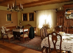 Zdjęcie przedstawia umeblowany pokój. Na pierwszym planie stoi nakryty do posiłku stół, a przy nim krzesła. Za stołem widoczny jest drewniany kredens. Po lewej stronie stoi mniejszy stolik i krzesła