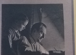 Zdjęcie przedstawia siedzącego mężczyznę, pochylonego nad biurkiem i stojącą za jego plecami dziewczynkę