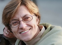 Zdjęcie przedstawia siedzącą uśmiechniętą kobietę w okularach