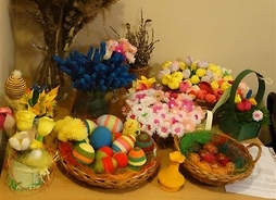 Zdjęcie przedstawia wiklinowe koszyczki wypełnione pisankami i kwiatami z bibuły. W szklanych wazonach stoją bazie oraz kwiaty z bibuły