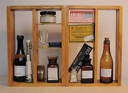 Zdjęcie przedstawia drewnianą półkę z pionowymi przegródkami, w których umieszczone są butelki, flakoniki, słoiczki