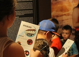 Grupa dzieci podczas zajęć w muzeum (zdjęcie wykonane z boku). Na pierwszym planie dziewczynka, która trzyma w jednej dłoni folder muzeum w Płocku, a w drugiej ołówek