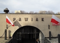 Zdjęcie przedstawia murowaną bramę więzienia Pawiak. Po obu jej stronach wiszą biało-czerwone flagi