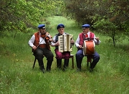 Zdjęcie w plenerze. Trzej muzykańci w ludowych strojach siedzą z instrumentami muzycznymi.