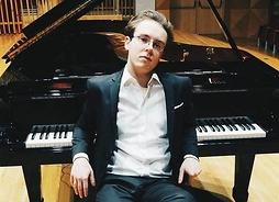 Młody mężczyzna w okularach i w stroju oficjalnym siedzi oparty o klawisze fortepianu