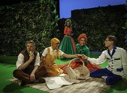 Zdjęcie z przedstawienia. Przedstawia grupę aktorów w kostiumach scenicznych siedzących na kocu. Przed nimi stoi wiklinowy kosz.