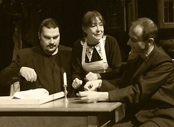 Troje aktorów podczas przedstawienia. Pośród dwóch mężczyzn przy stole siedzi kobieta