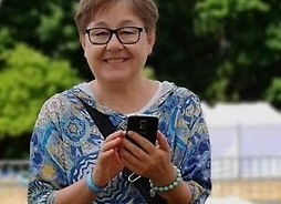 Zdjęcie portretowe Emilii Pituchy. Uśmiechnięta kobieta w okularach, w dłoniach trzyma telefon komórkowy, zdjęcie w plenerze