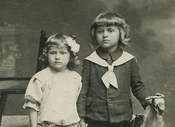 Zdjęcie archiwalne, pozowane, przedstawia dwójkę kilkuletnich dzieci. Chłopczyk trzyma dłoń na głowie drewnianego konia na biegunach, dziewczynka trzyma w ręku różę