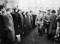 Zdjęcie archiwalne z wizytacji Pawiaka przez Himmlera. Przedstawia grupę niemieckich żołnierzy w mundurach, po jednej stronie, naprzeciwko nich w szeregu stoją mężczyźni w cywilnych ubraniach.