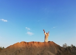 Zdjęcie w plenerze. Mężczyzna odwrócony tyłem, klęczy na wysokiej górze usypanej z piachu, z rękami uniesionymi ku niebu.