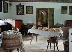 Wnętrze wiejskiej chaty. Na pierwszym planie drewniana balia na pranie, w głębi drewniana ława, na której leżą bochenki chleba