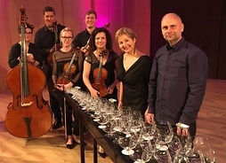 Grupa artystów w strojach oficjalnych z instrumentami muzycznymi. Przed nimi stoi stół zastawiony szklanymi kieliszkami wykorzystywanymi do gry.