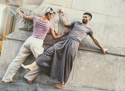 Tancerze - mężczyzna i kobieta stojący na gzymsie budynku. Oboje trzymają się szczelin w kamiennej ścianie.
