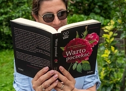Kobieta w okularach trzymająca przed sobą otwartą książkę. Zdjęcie w plenerze.