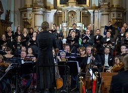 Na pierwszym planie dyrygent odwrócony plecami do widzów, dyruguje grupą muzyków siedzących na krzesłach i grających na instrumentach. Za nimi stoją chórzyści. Zdjęcie wykonane w kościele.