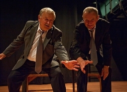 Dwóch mężczyzn w garniturach podczas występu na scenie. Obaj jednocześnie schylają się po coś, wyciągając dłonie.