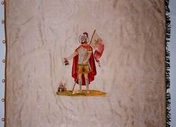 Sztandar z wizerunkiem św. Floriana. Mężczyzna w stroju rzymianina, w płaszczu i hełmnie na głowie i horągwią w jednej ręce, w drugiej trzyma naczynie z wodą, którą gasi pożar domu.