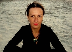 Kobieta z umpiętymi z styłu włosami siedzi na piasku. Zdjęcie do pasa.