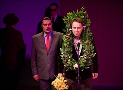 Dwaj aktorzy w garniturach podczas odgrywania sceny przedstawienia, jeden z nich na szyi zawieszony dużych rozmiarów wieniec z liści i kwiatów, a w ręku trzyma bukiet kwiatów.