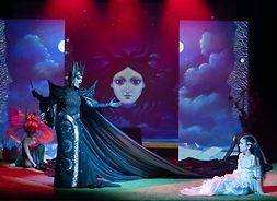 Scena z opery. Artystka w kostiumie scenicznym stoi na scenie i wykonuje arię operową, zwrócona w stronę artystki, która siedzi na podłodze sceny.