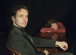 Mężczyzna w garniturze siedzi przy stole z jedną ręką położoną na blacie. Pod drugiej stronie stołu siedzi kobieta, ma głowę położoną na ramionach, ułożonych na blacie stołu.