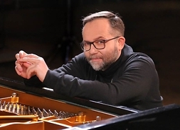Mężczyzna w okularach siedzący przy fortepianie z rękoma wspartymi na obudowie