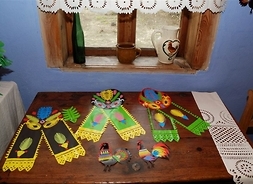 Papierowe wycinanki ułożone na drewnianym stole ustawionym przy oknie ozdobionym wycianą z papieru firanką. Na parapecie stoi od lewej: butelka, gliniany kubek i dzbanek.