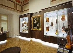 Sala wystawowa muzeum. Gabloty ze strunowymi instrumentami muzycznymi, wiszące na ścianie, pośrodku gablot wisi portret kobiety w długiej sukni i harfą w tle..