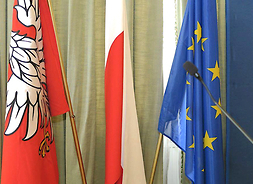 W zbliżeniu trzy flagi w sali obrad Sejmiku, od lewej flaga Mazowsza, flaga Polski, flaga Unii Europejskiej.