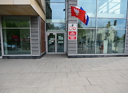 Widok na przeszklone drzwi frontowe, nad wejsciem wiszą flagi UE, Polski i Mazowsza